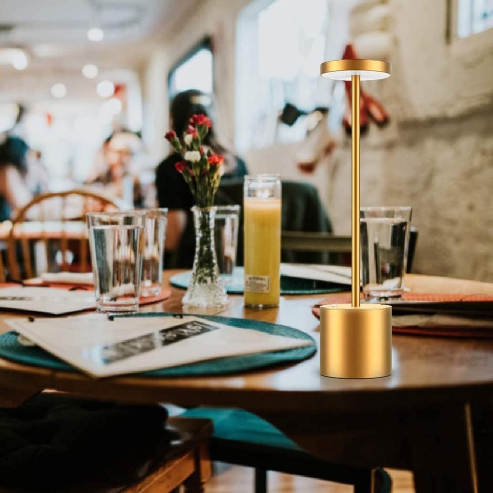 Eine goldene Tischlampe auf einem Holztisch. Ein Blumenarrangement, Gläser und ein Saftglas sind ebenfalls vorhanden.