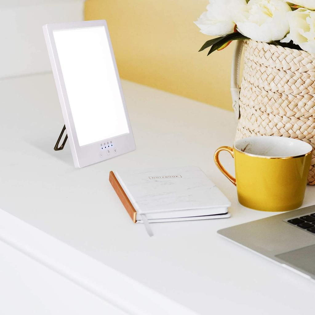 Eine weiße Lichttherapielampe, ein Notizbuch, ein gelber Becher, ein Laptop und ein Korb mit Blumen auf einem Schreibtisch.