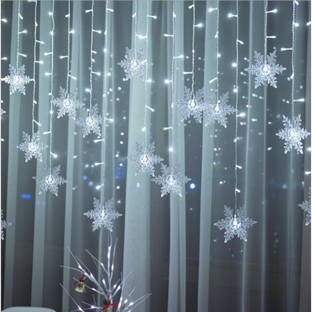 Schneeflockenlichter hängen vor einem transparenten Vorhang. Weiße Lichter und ein kleiner Baum.