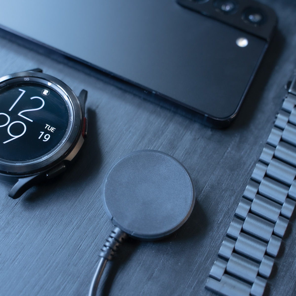 Schwarze Smartwatch und Telefon auf einem Tisch, mit Ladepad und Armband.
