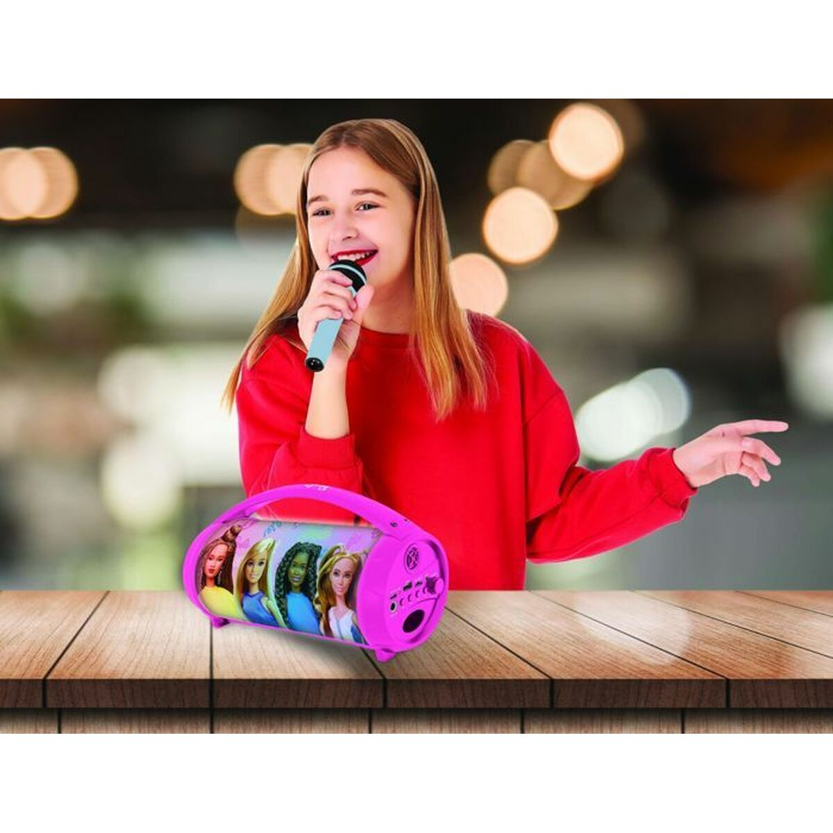 Ein Mädchen singt in ein Mikrofon, ein pinkfarbener Barbie-Lautsprecher auf einem Holztisch.