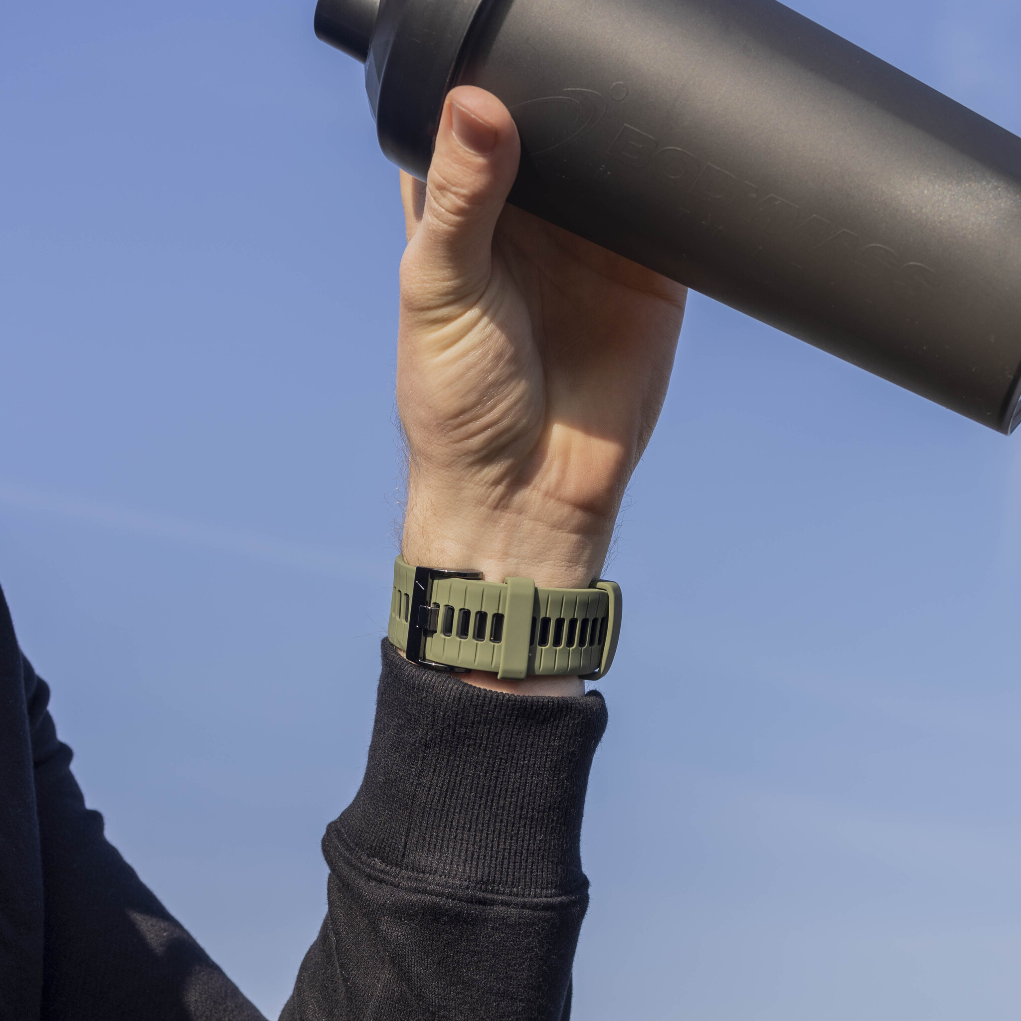Das Handgelenk einer Person mit einer grünen Uhr und einer schwarzen Shaker-Flasche in der Hand.