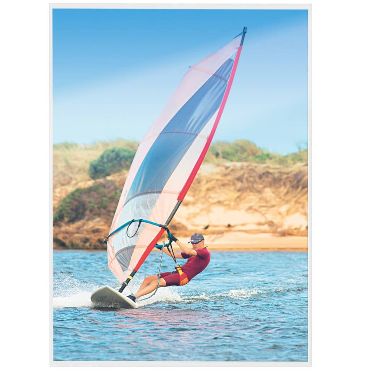Windsurfer auf blauem Wasser, mit blauem Himmel und Sandstrand im Hintergrund.