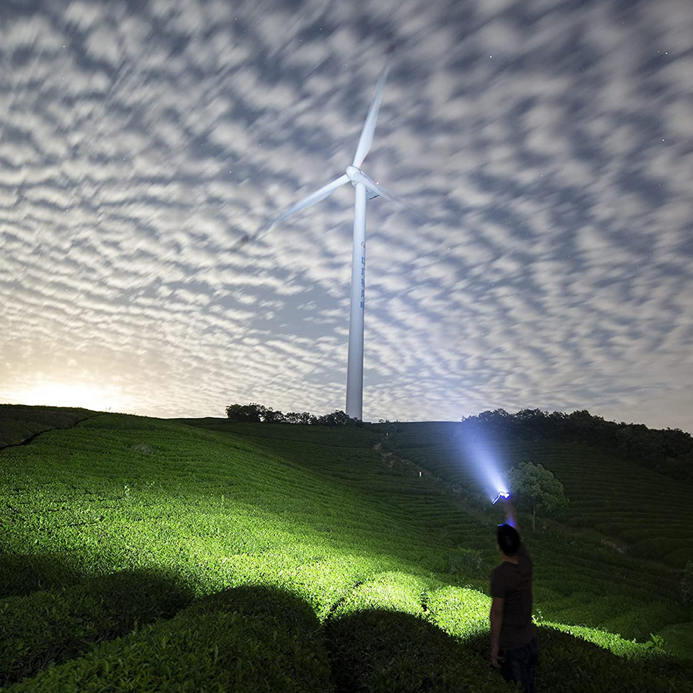 Eine Person hält nachts eine Taschenlampe und beleuchtet eine Windturbine auf einem grünen Hügel.
