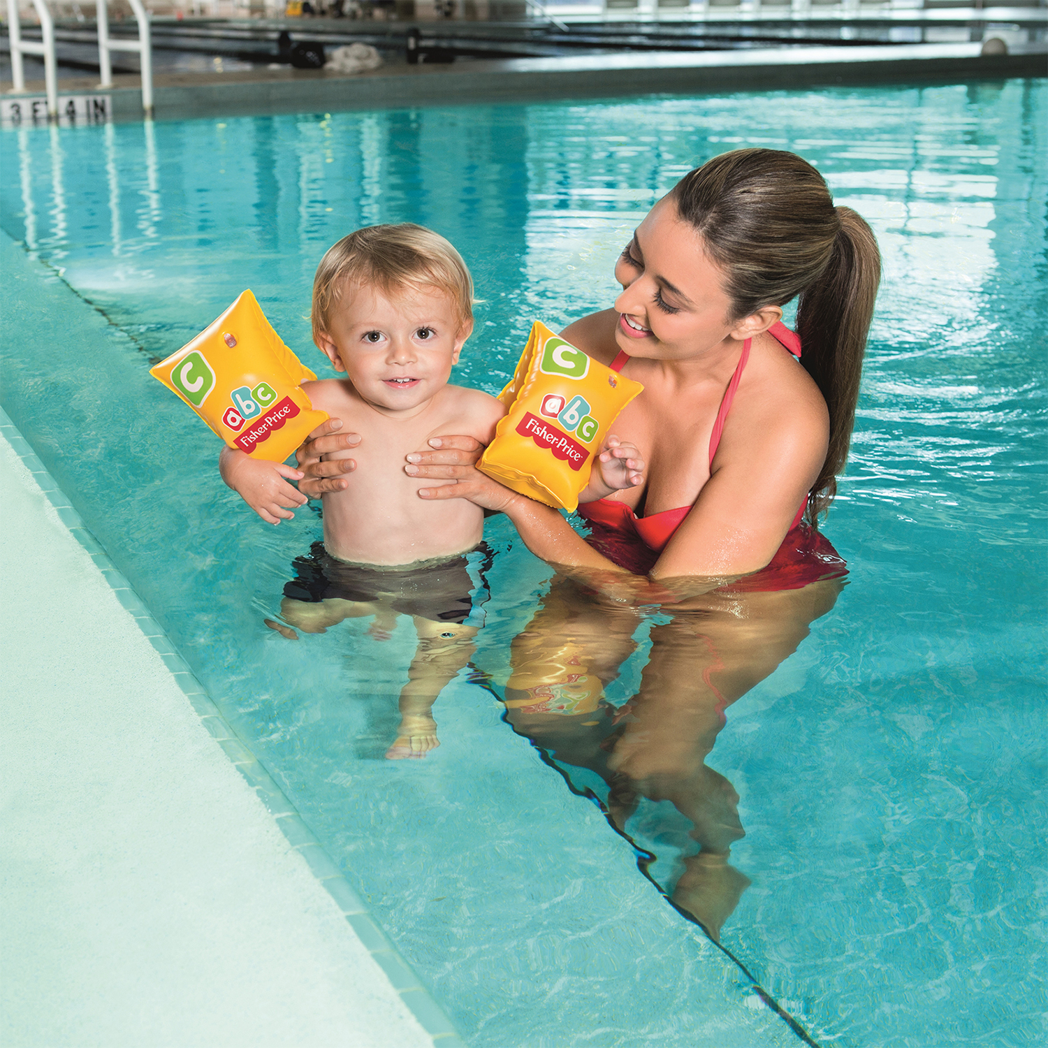 Una mujer y un niño están en una piscina. El niño sostiene flotadores de brazo. Están sonriendo a la cámara.
