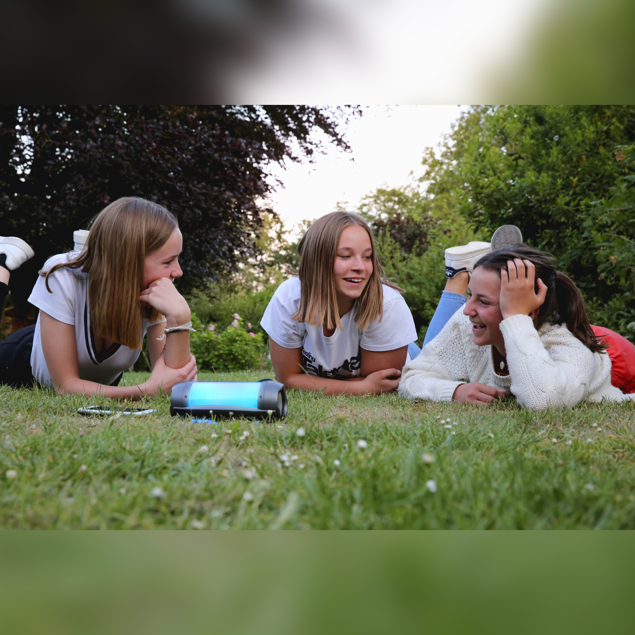 Drie jonge vrouwen die op het gras liggen te lachen, een speaker staat naast hen.