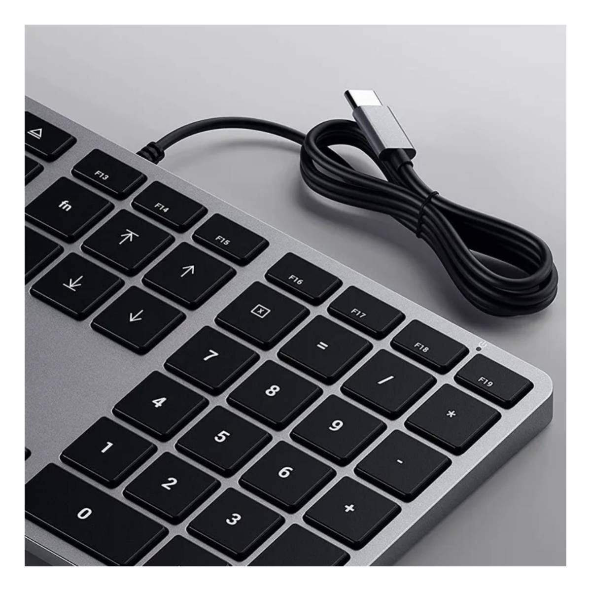 Un teclado plateado con teclas negras y un cable USB-C conectado, sobre una superficie gris.