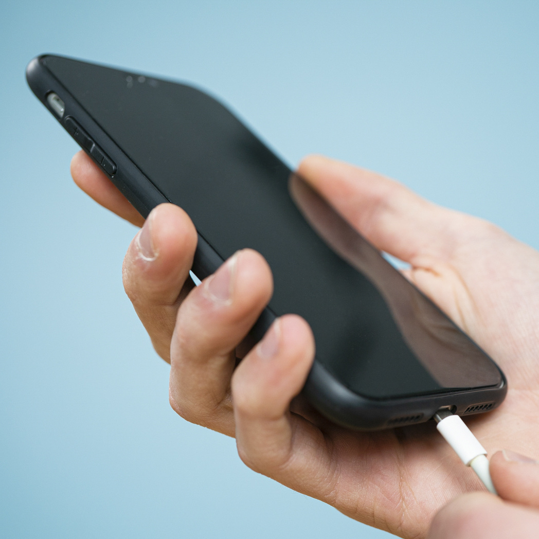 Una mano che tiene un telefono nero con un caricabatterie bianco collegato, su sfondo blu.
