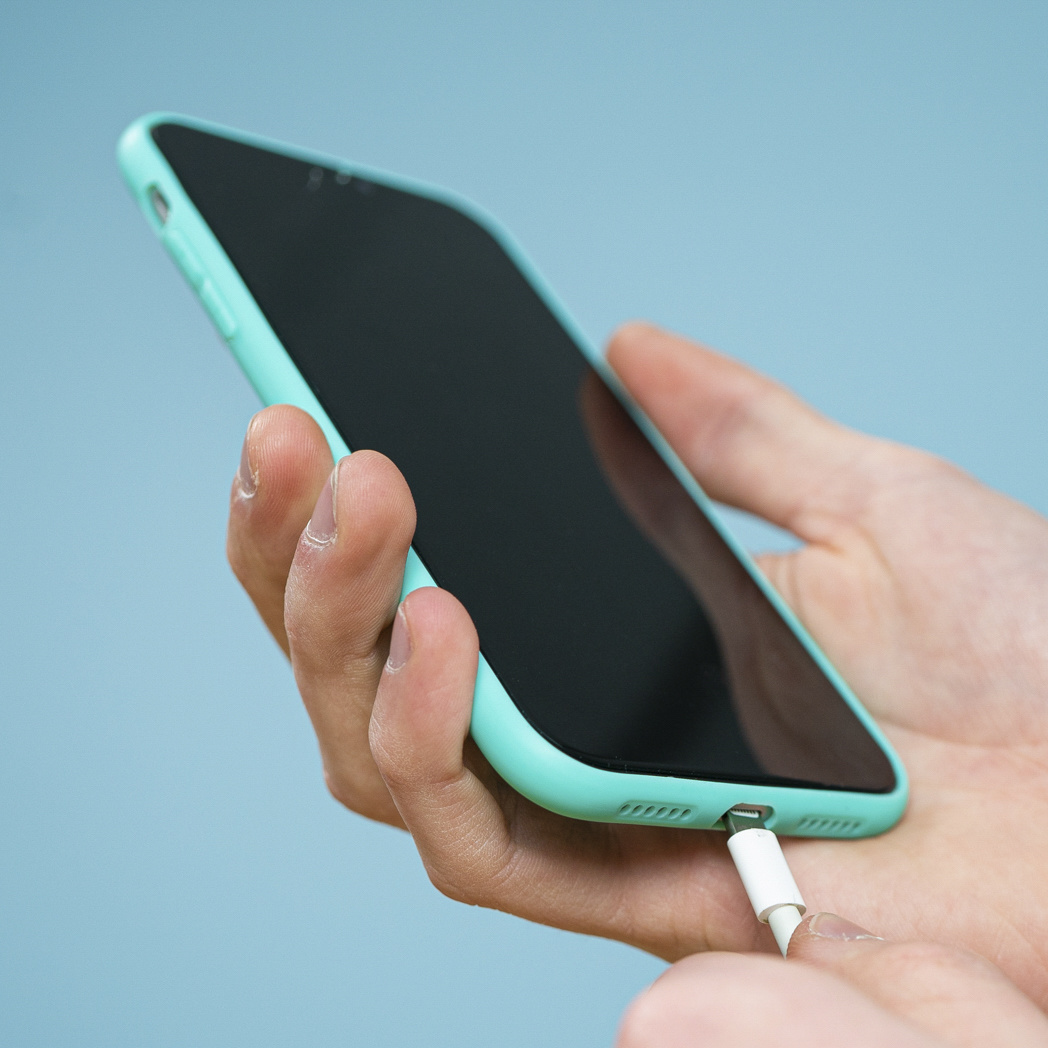 Hand die een mintgroene telefoon vasthoudt met een witte oplaadkabel aangesloten. De achtergrond is blauw.
