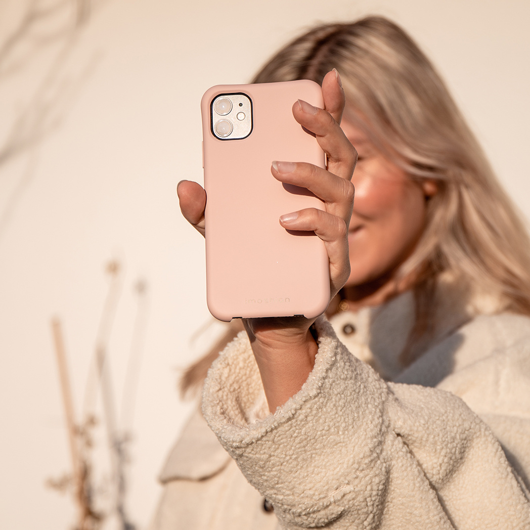 Blonde vrouw met een roze telefoonhoesje. Ze draagt een witte jas. Achtergrond is een muur.
