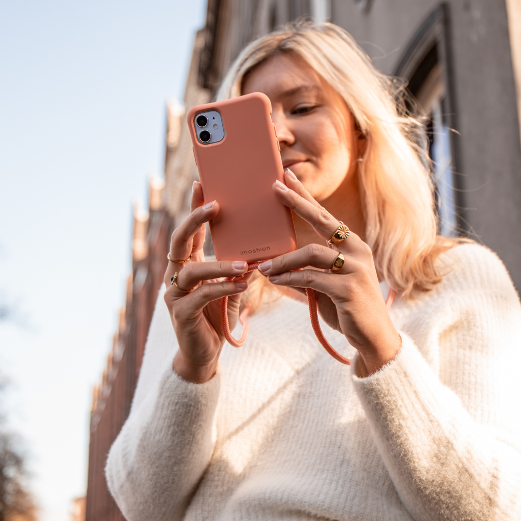 Blonde vrouw die een koraalroze telefoonhoes vasthoudt. Ze draagt ringen en een witte trui; een gebouw op de achtergrond.