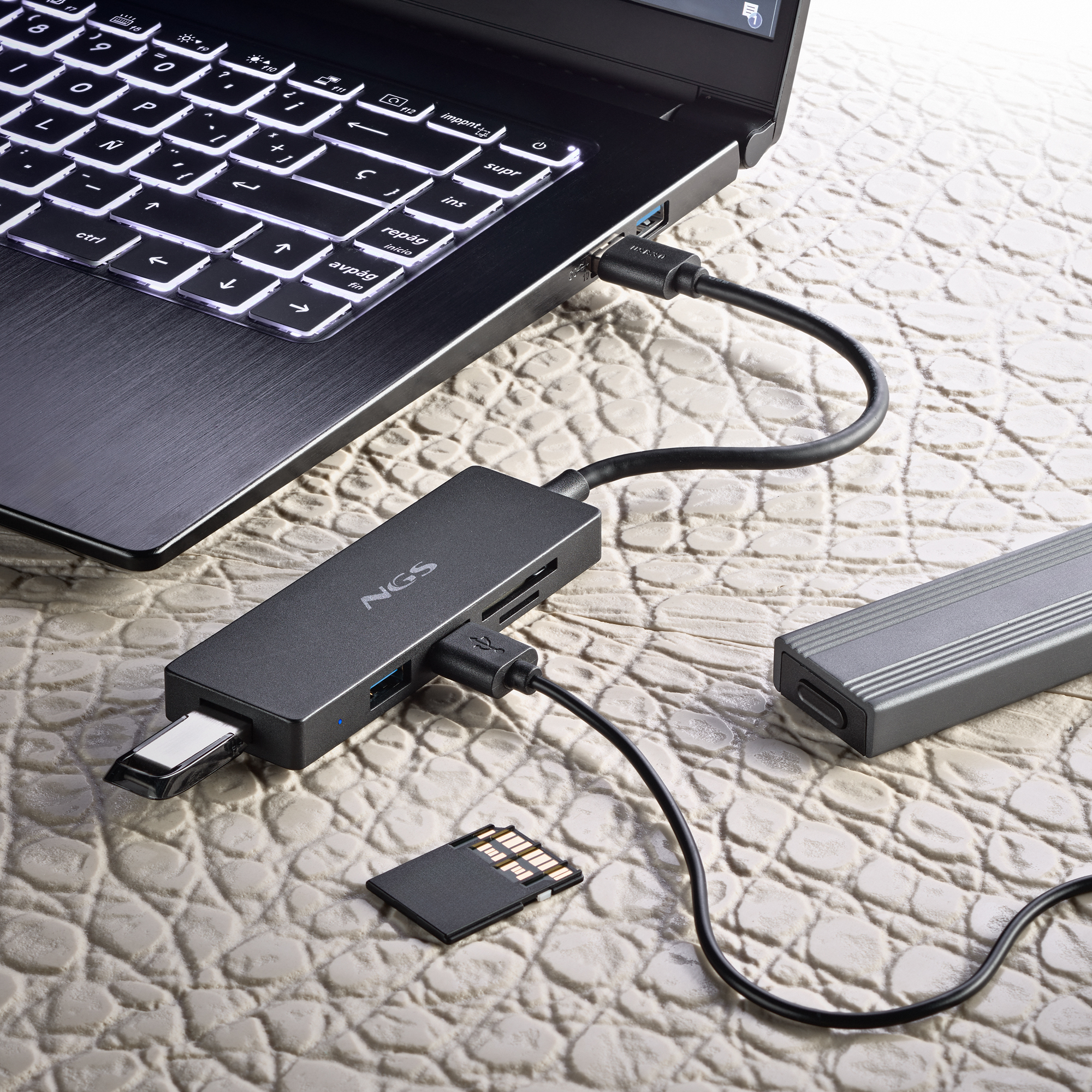 Laptop, verbunden mit einem Hub mit SD-Karte, USB- und Stromkabeln auf strukturierter Oberfläche.