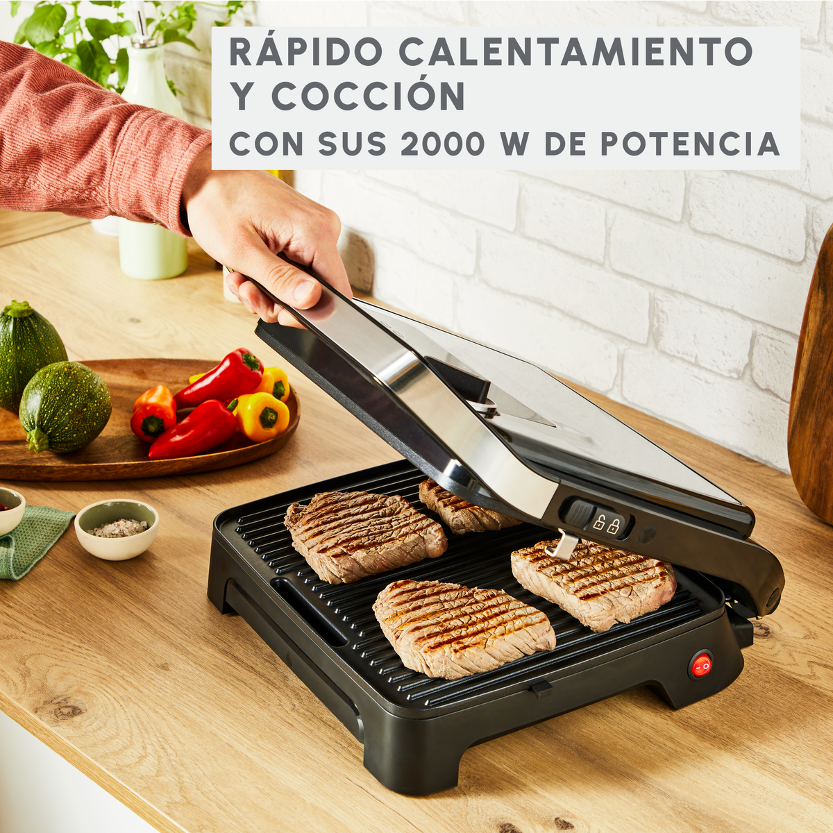 Grill z gotującymi się stekami, z ręką osoby, która go otwiera. Widać warzywa i ceglany mur.