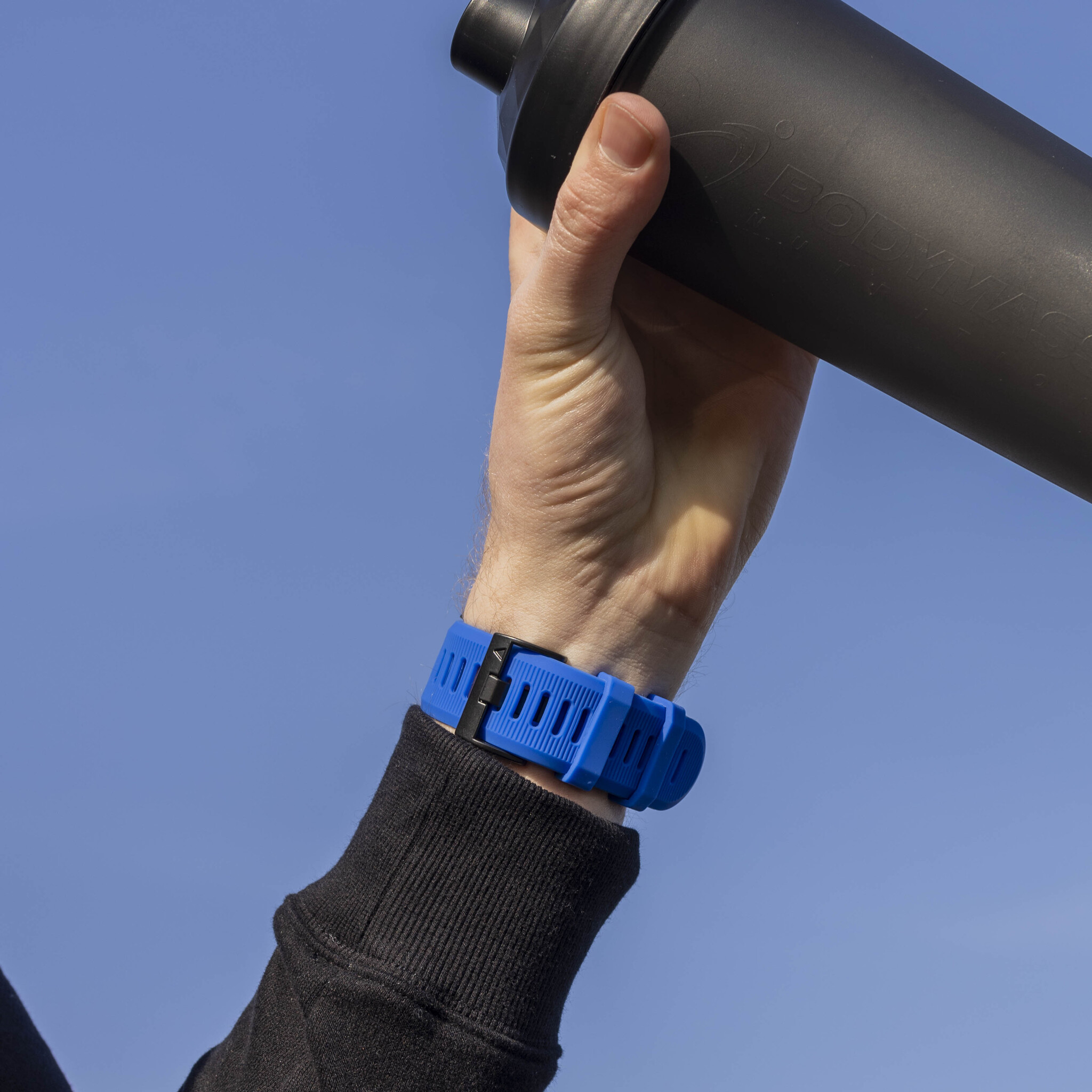 Das Handgelenk einer Person mit einem blauen Band und einem schwarzen Getränkemixer in der Hand.