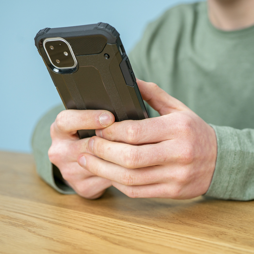 Mani di una persona che tengono un telefono nero in una custodia protettiva su un tavolo di legno.
