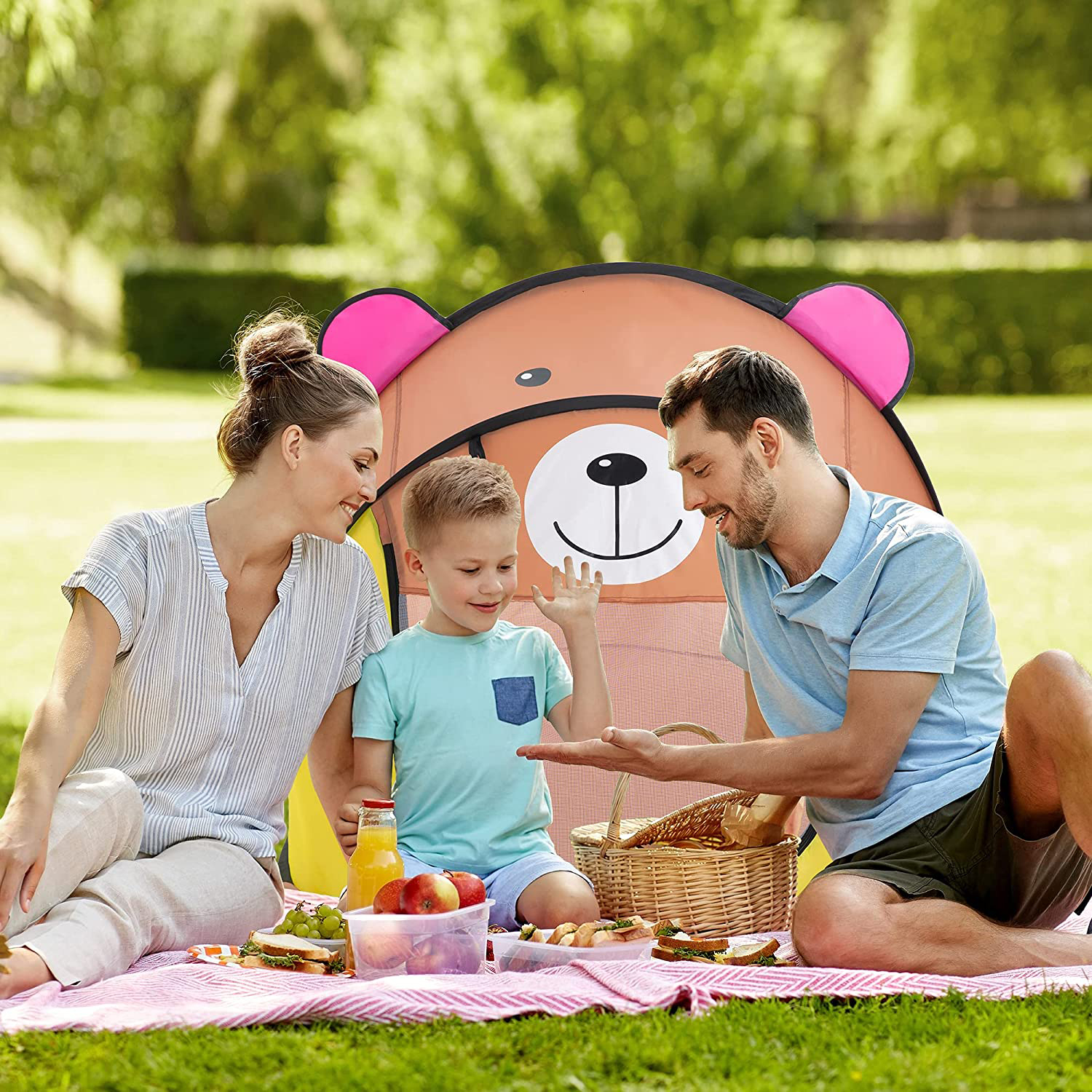 Picnic familiar con manta rosa, tienda de oso, comida y fondo soleado.
