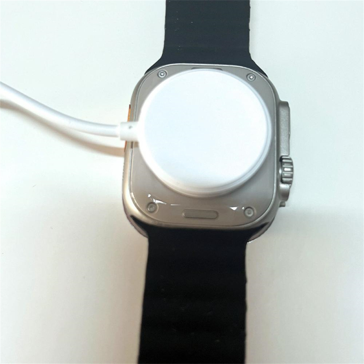 Eine silberne Apple Watch Ultra mit angeschlossener Ladescheibe und einem schwarzen Armband. Auf weißem Hintergrund.