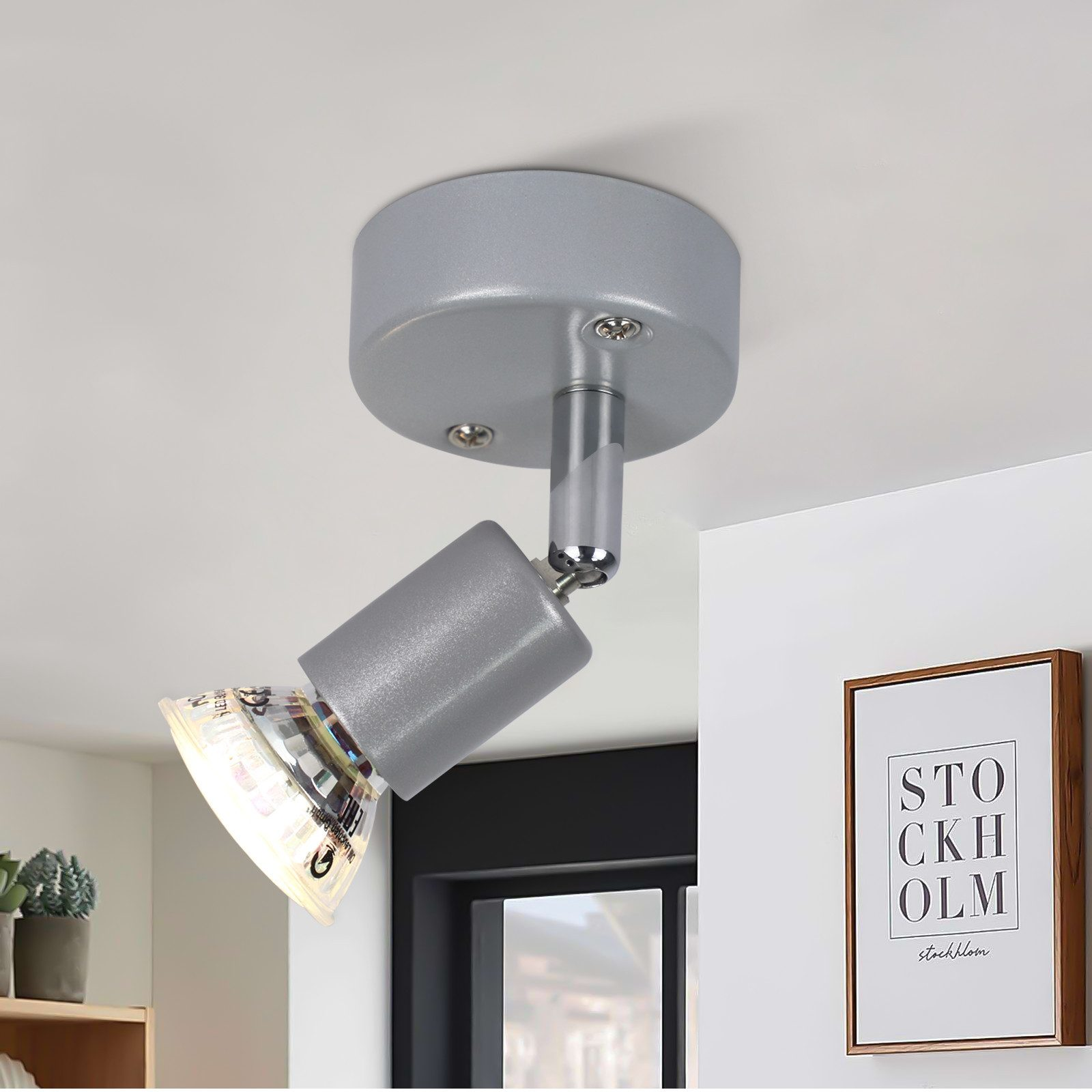 Szary reflektor sufitowy. Ma cylindryczny korpus i skierowany jest w dół. Po prawej stronie znajduje się oprawiony obraz.