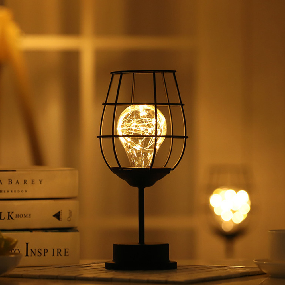 Schwarze Metalllampe mit Glühbirne, die einem Weinglas ähnelt, auf einem Tisch. Andere Lampe im Hintergrund.