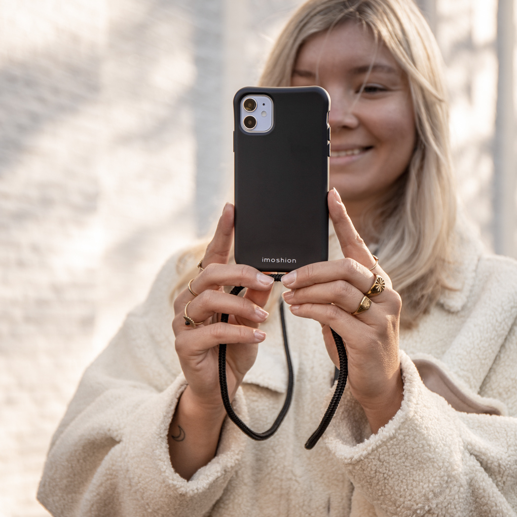Een vrouw die een zwarte telefoon vasthoudt met een hoesje en een koord, lachend naar de camera.