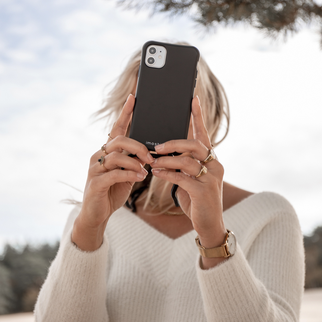 Handen van een vrouw die een telefoon vasthouden om een foto te maken. Ze draagt een witte trui en ringen.