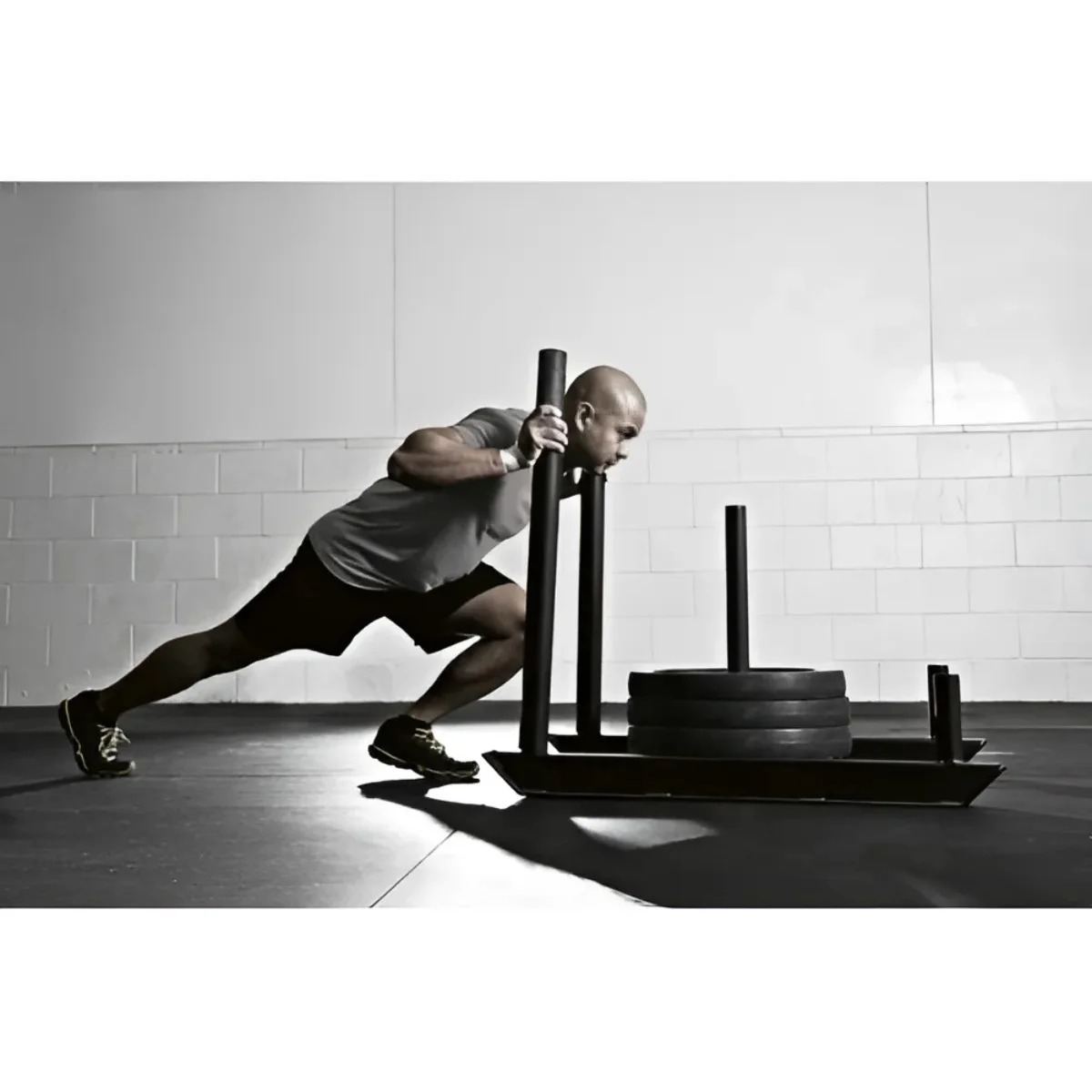 A man pushing a weighted sled in a gym. He is wearing black shorts and a gray shirt.