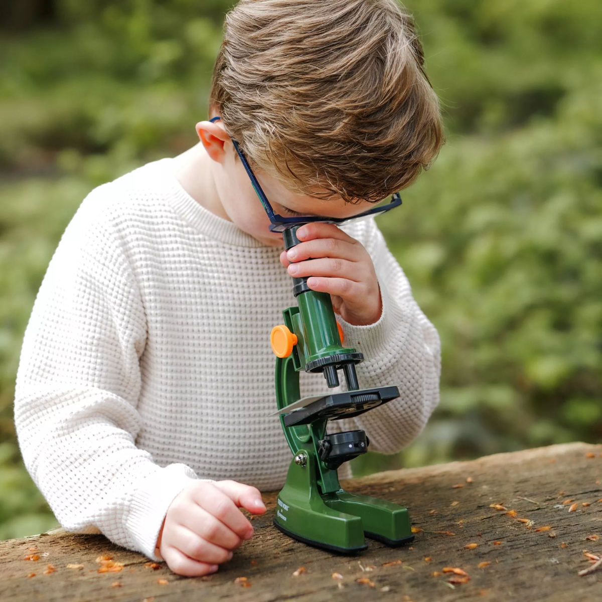 Un niño mirando a través de un microscopio verde sobre una superficie de madera, al aire libre.