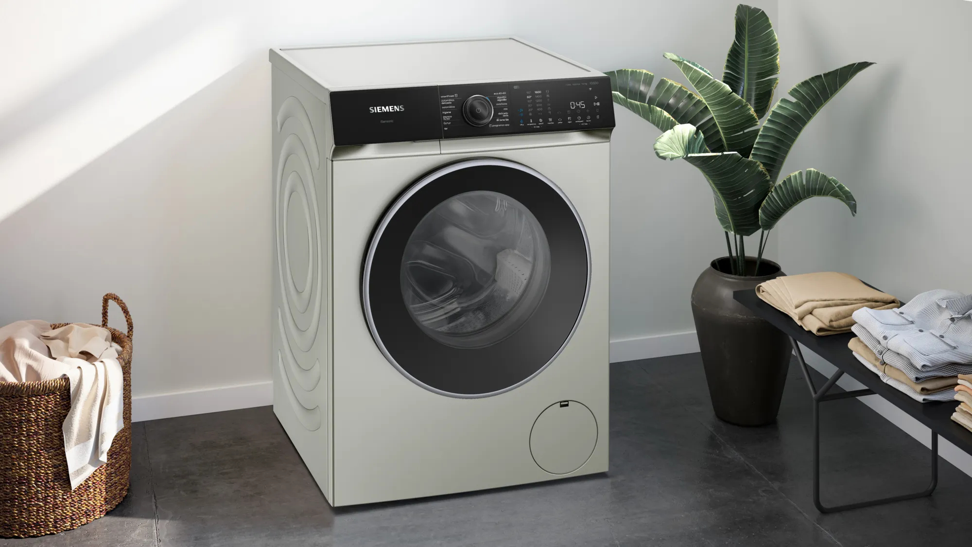 Una lavadora Siemens de color plateado. Está junto a una cesta con ropa y una planta sobre un fondo oscuro.