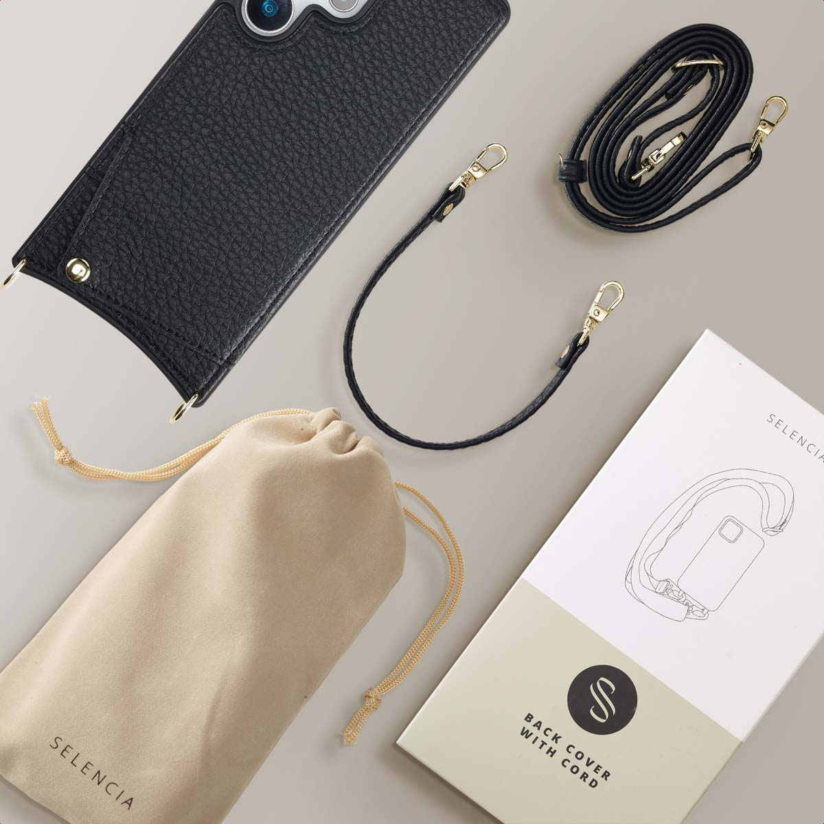 Custodia per telefono nera con cinturini neri e dorati, una borsa beige e una scatola bianca. Superficie grigia.