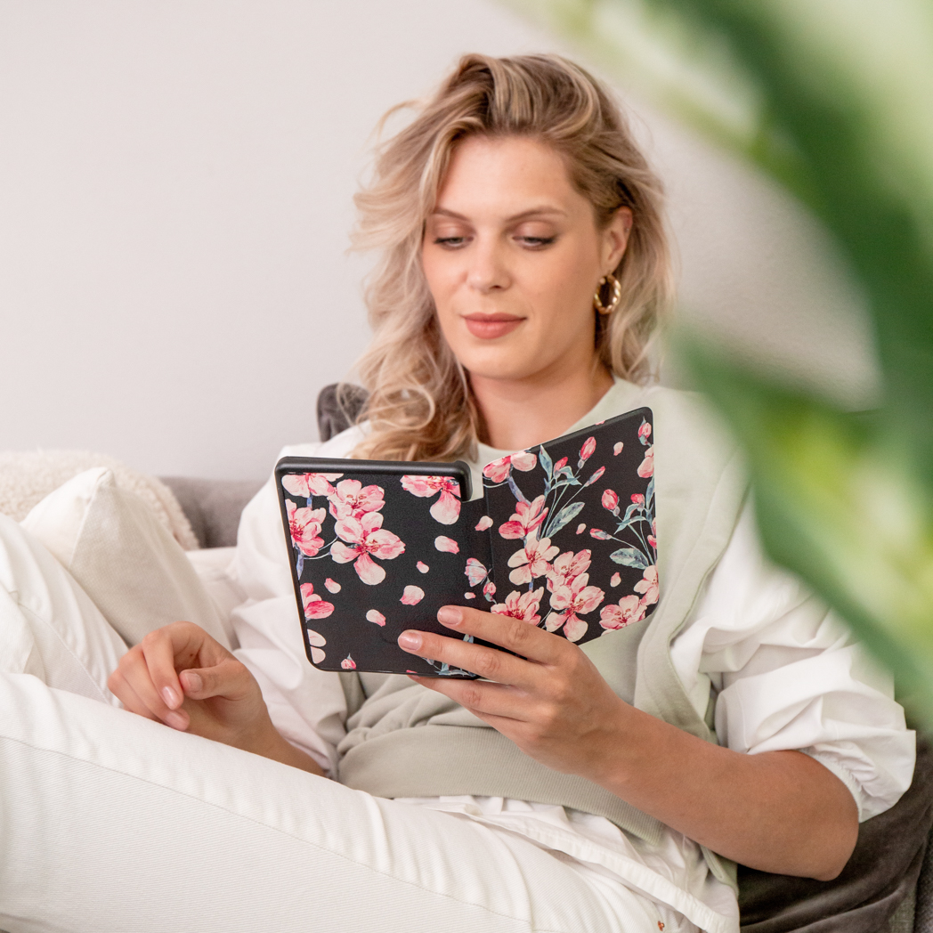 Femme lisant un livre avec un étui floral, assise sur un canapé. Une plante verte à droite.