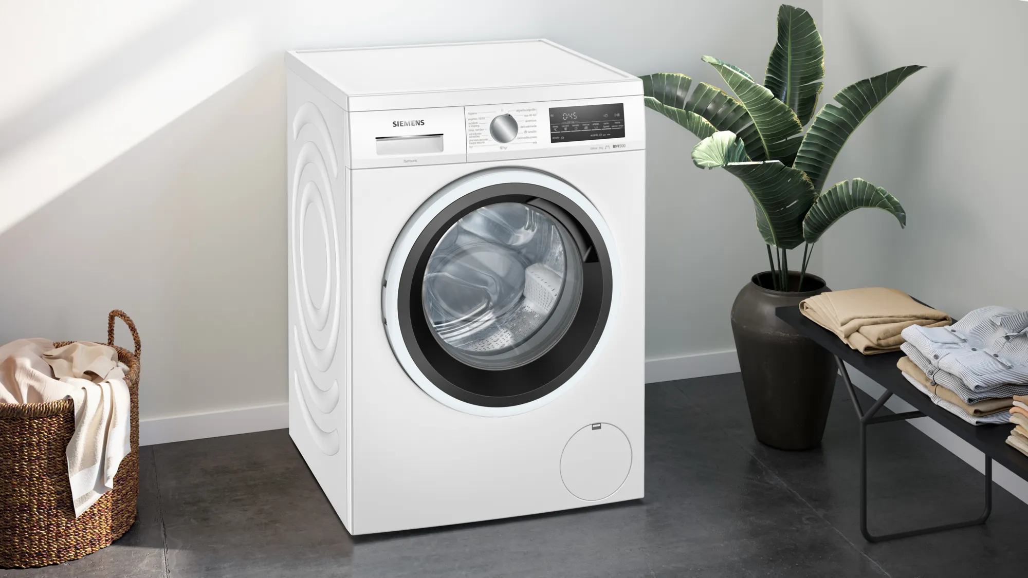 Una lavadora Siemens blanca en una habitación. Al lado, una planta en maceta, una cesta y ropa doblada.