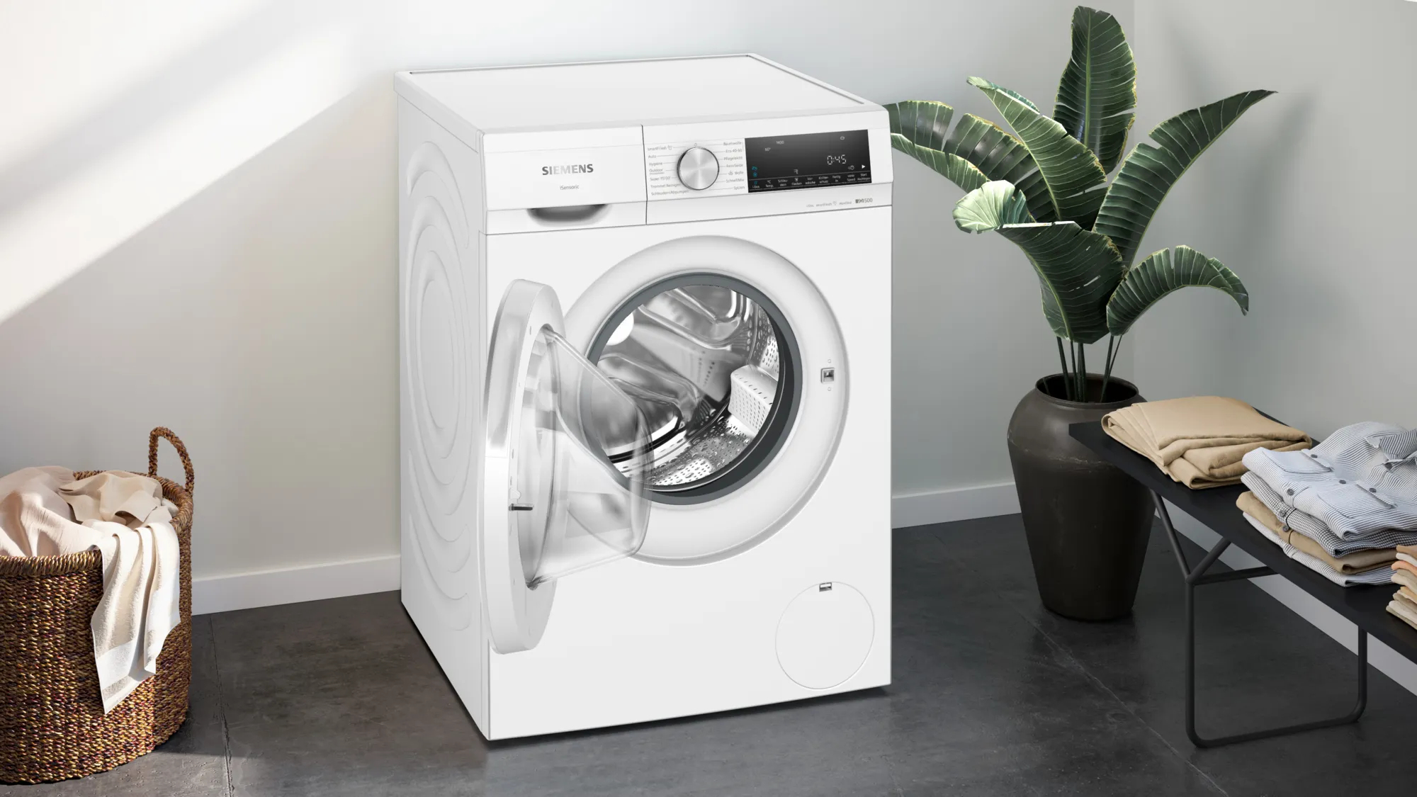 Weiße Siemens Waschmaschine mit offener Tür, neben einem Weidenkorb und einer Bank mit gefalteter Kleidung.