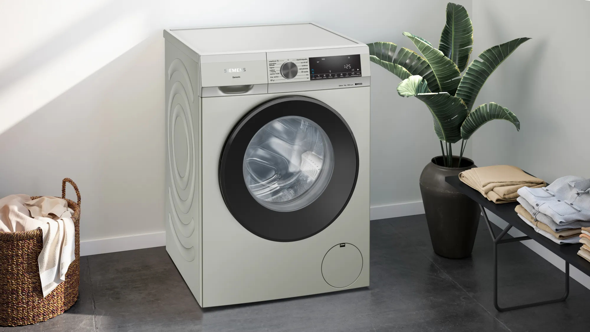 Una lavadora plateada con puerta negra. Una planta y una cesta con ropa a su lado.