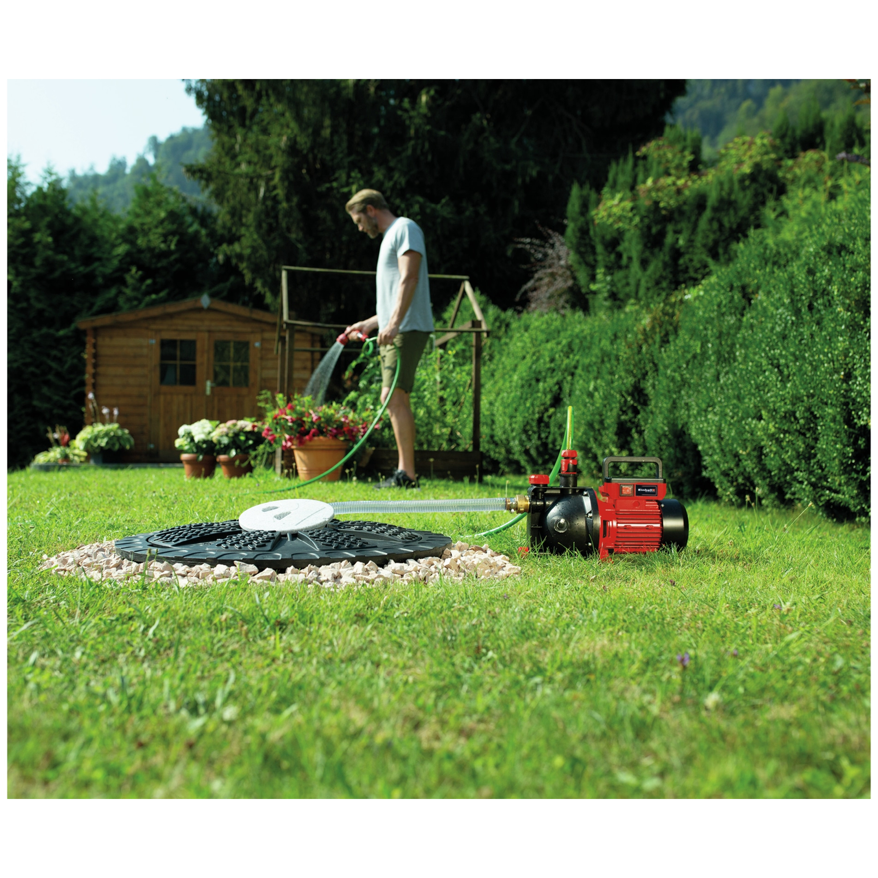 Mężczyzna podlewający rośliny w ogrodzie z czerwoną pompą wodną na pierwszym planie i drewnianą szopą w tle.