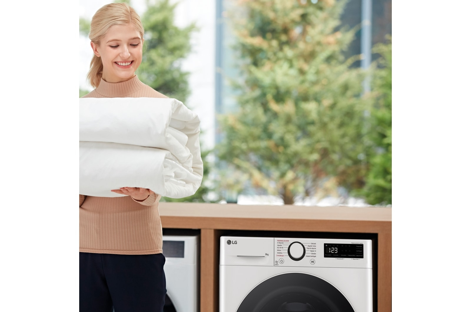 Kobieta trzyma złożoną białą pościel, obok pralki i okna.