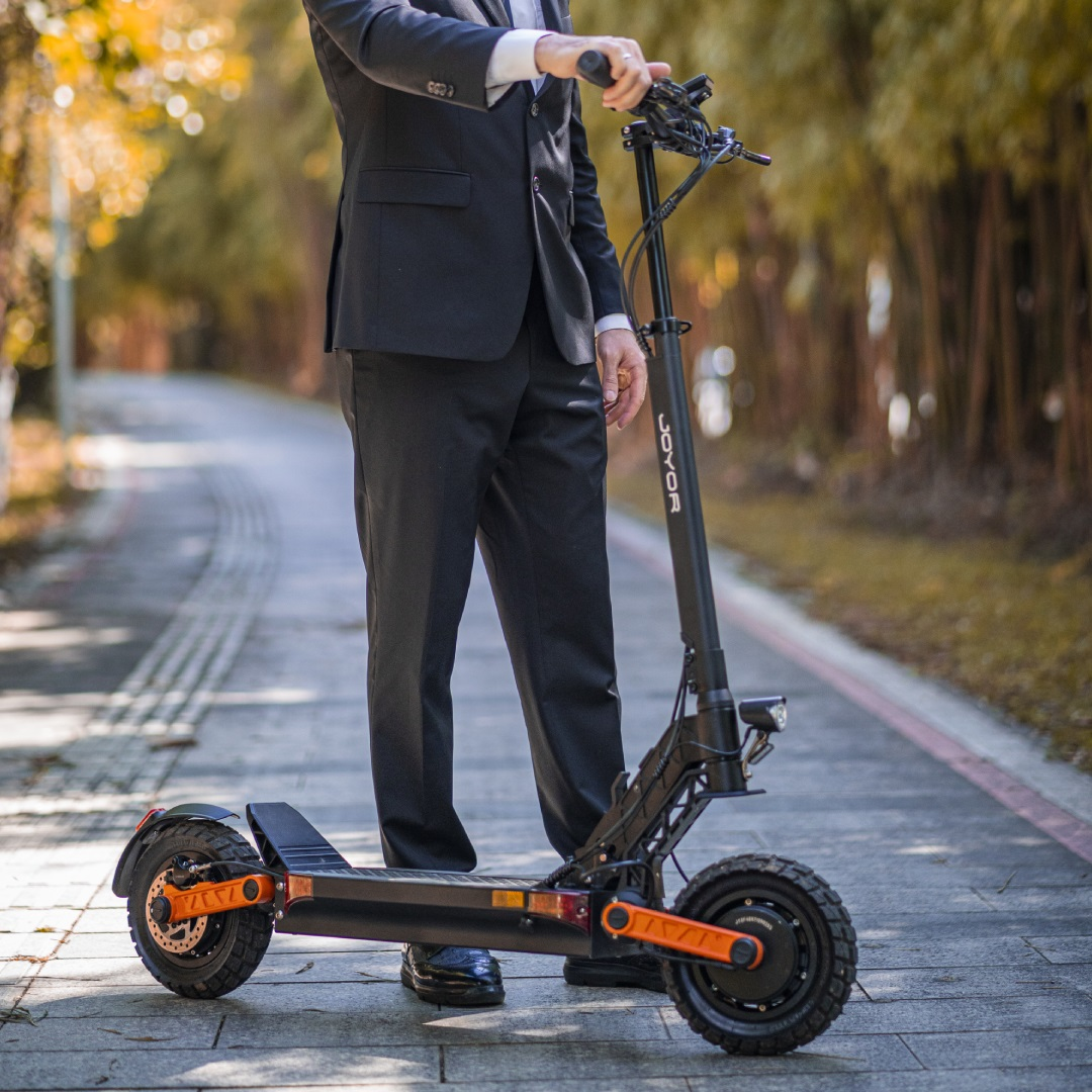 Ein Mann im Anzug steht neben einem schwarzen COCOOR Elektroroller auf einem Weg. Der Weg ist gepflastert.