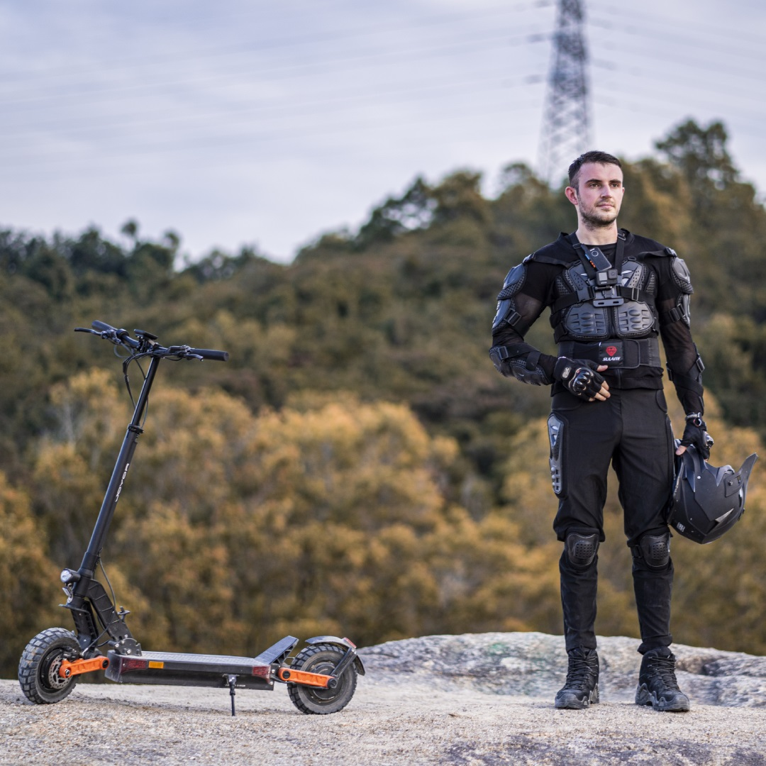 Mann in Schutzkleidung mit Elektroroller. Steht auf Felsen, Hintergrund mit Hügeln und Himmel.