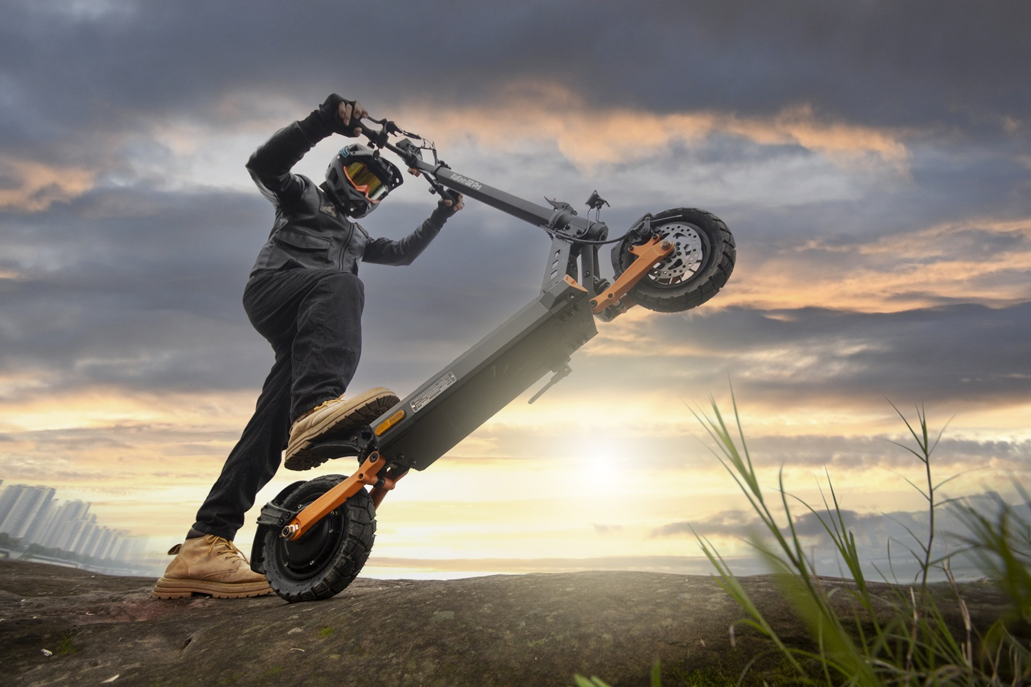 Uomo con casco su uno scooter che fa un'impennata, contro un tramonto.