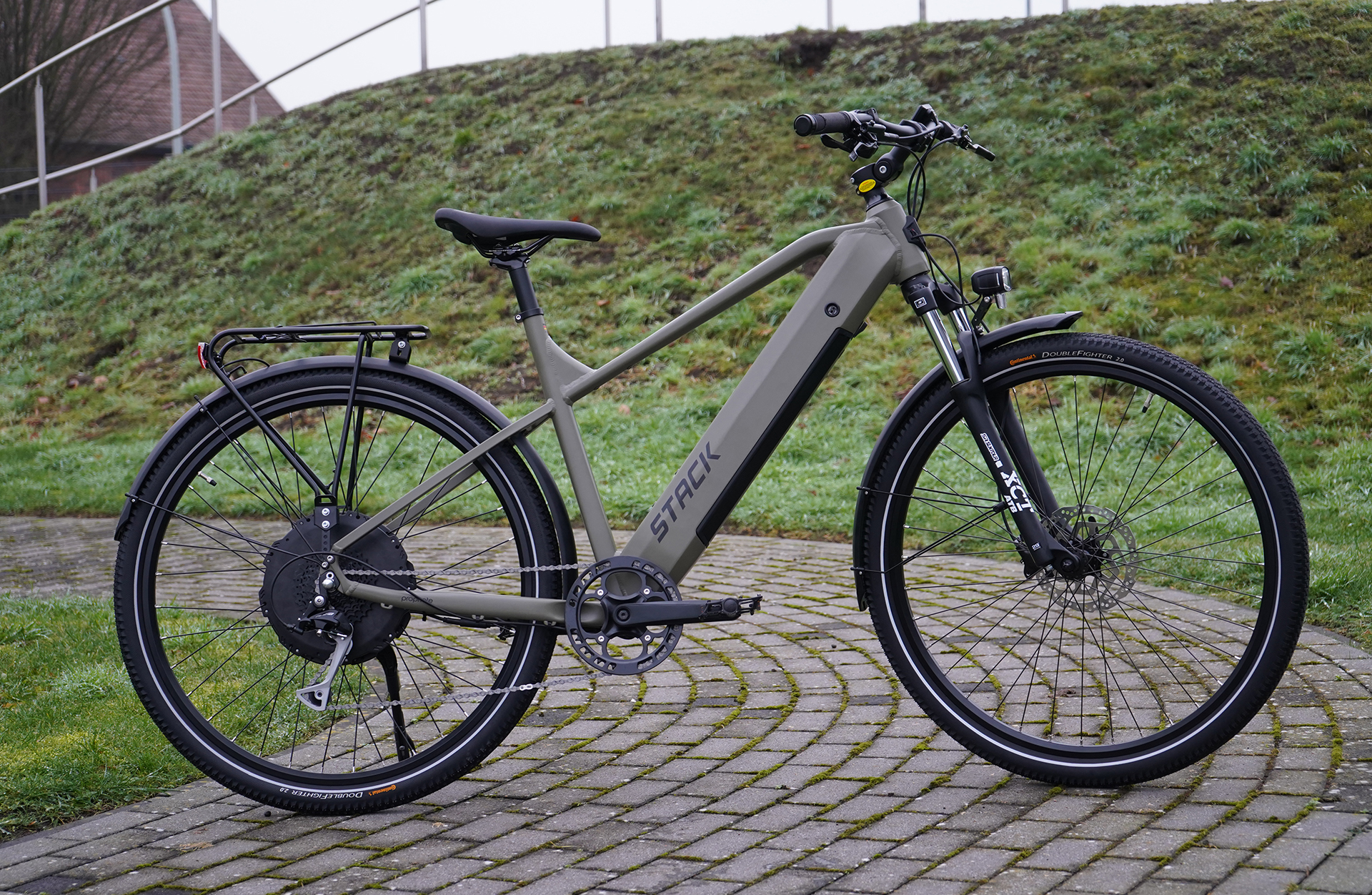 Ein olivgrünes Elektrofahrrad steht auf einem gepflasterten Weg, mit einem grasbewachsenen Hintergrund.