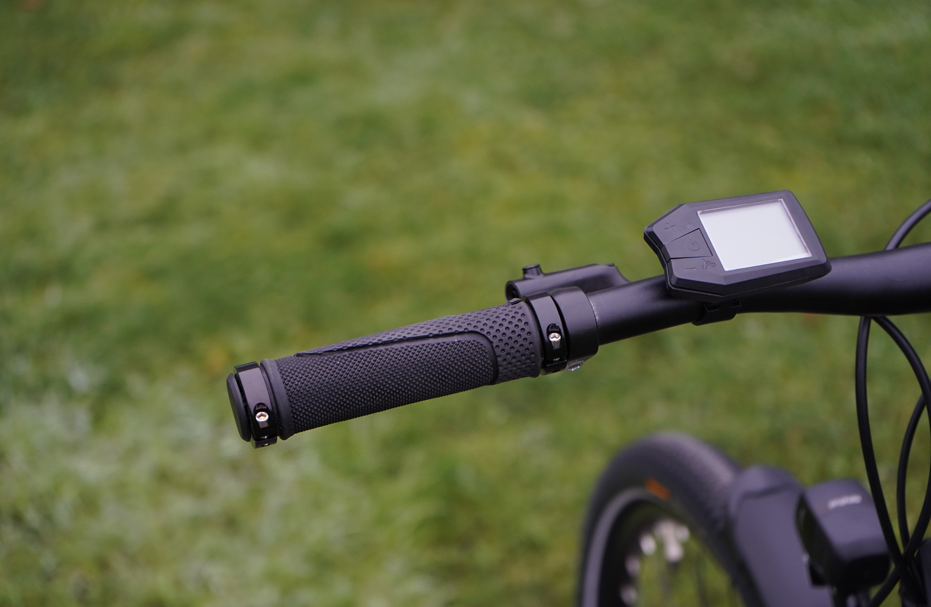 Fahrradlenker mit Display, schwarzer Griff. Verschwommenes Rad im Hintergrund, auf verschwommenem grünen Hintergrund.