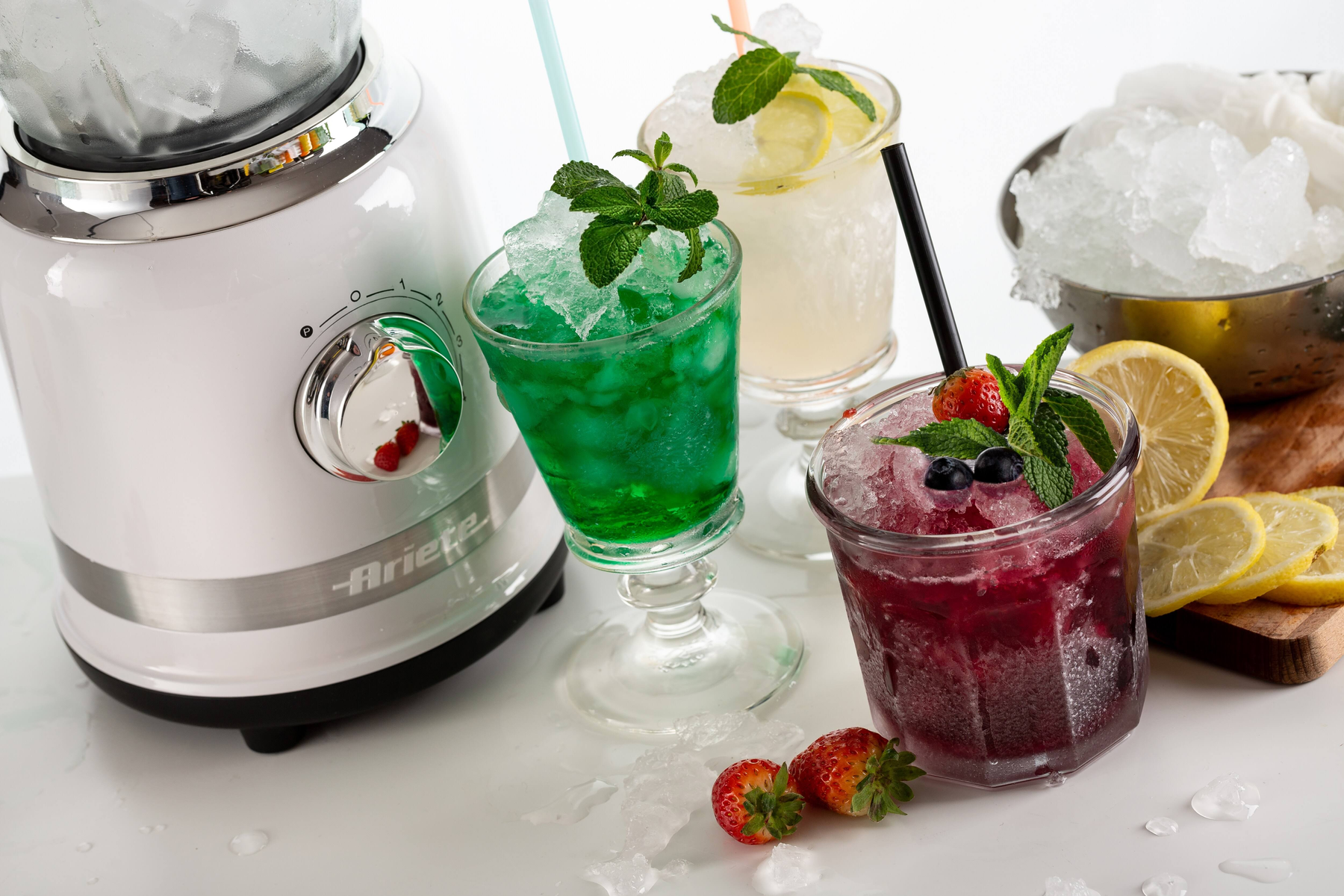 Drie cocktails met rietjes, munt en ijs, naast een blender en citroenen.
