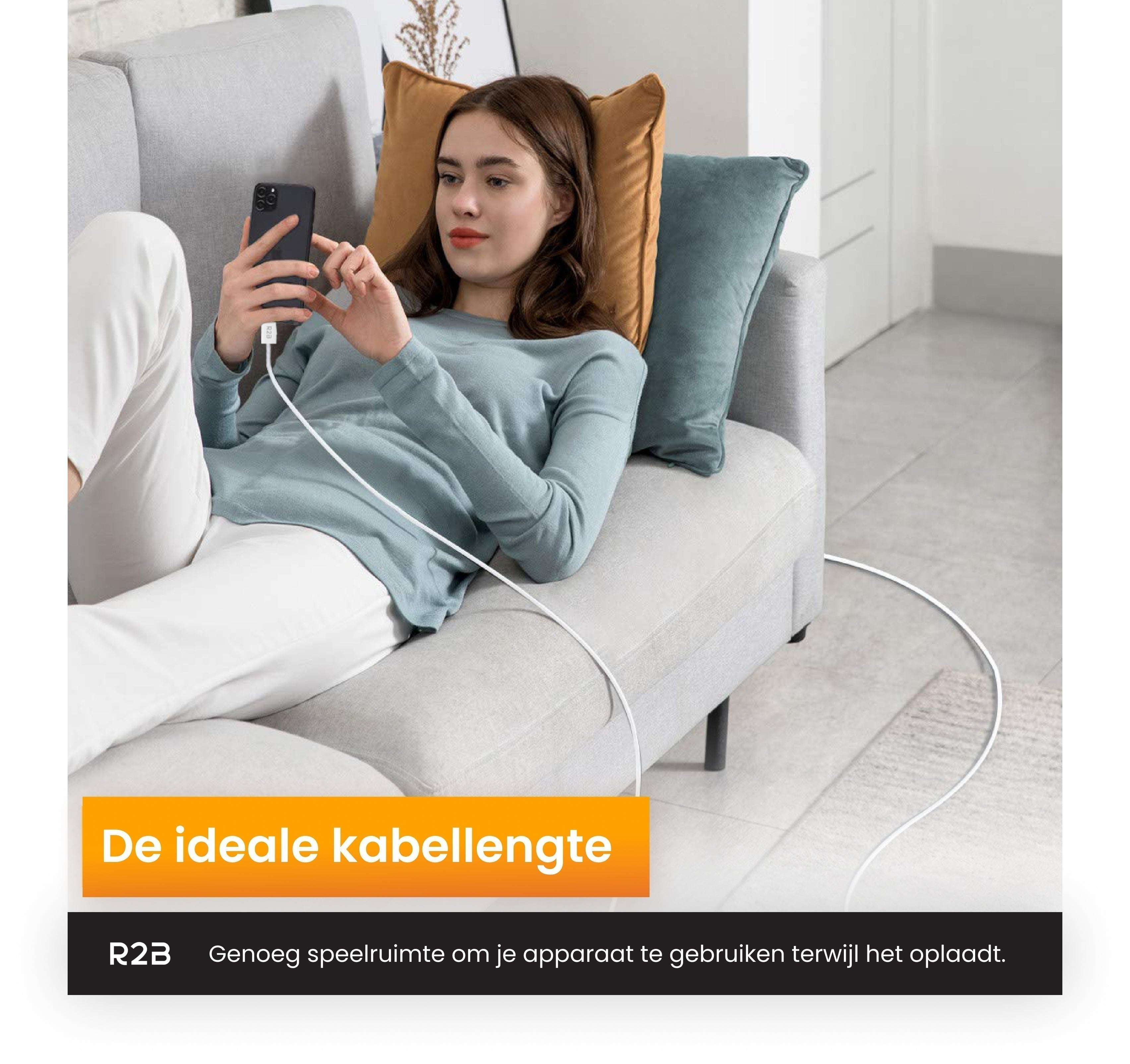 A woman sitting on a couch, holding a phone with a charging cable connected. Blue and orange pillows.