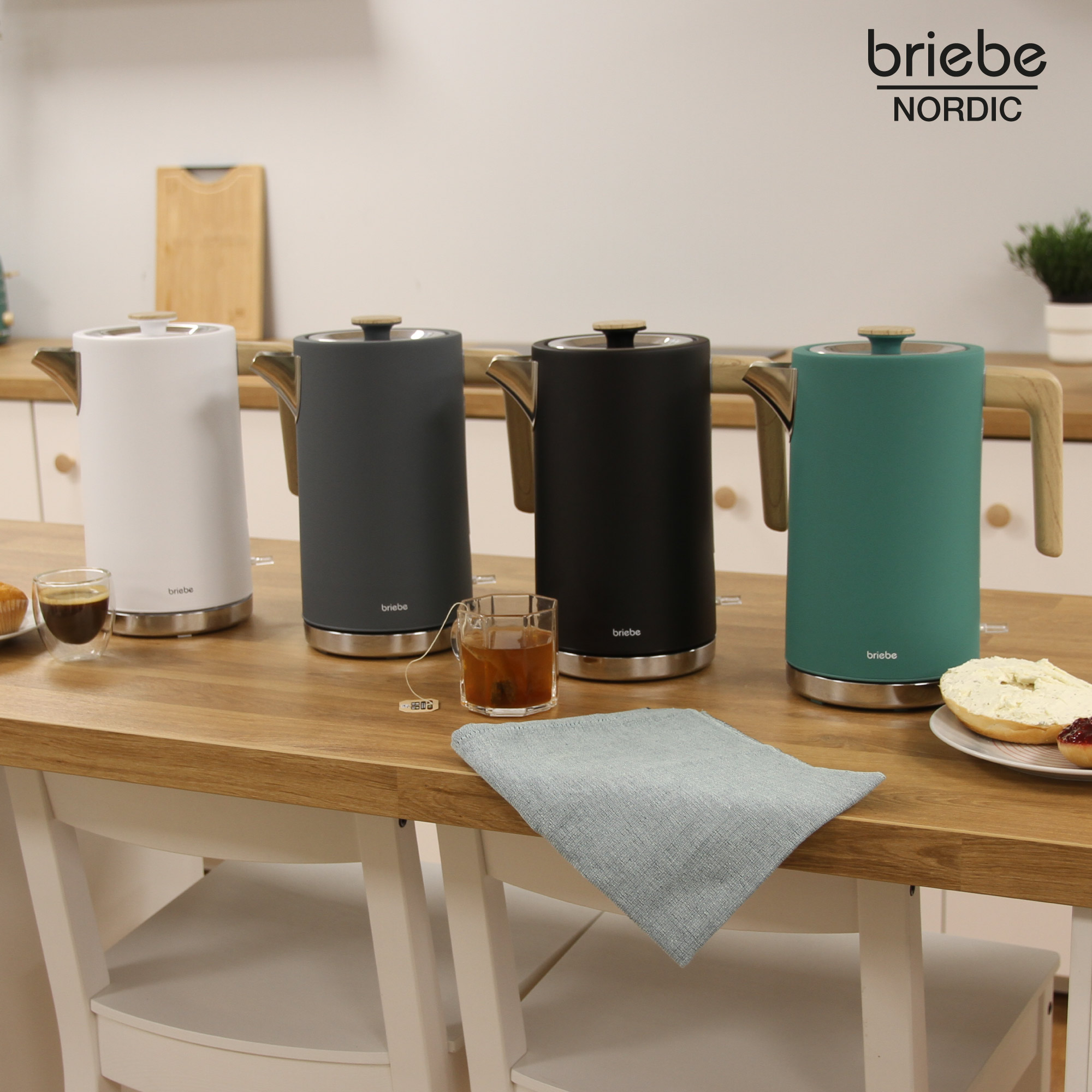 Cuatro hervidores de diferentes colores sobre una mesa de madera, con una taza de té y una servilleta.