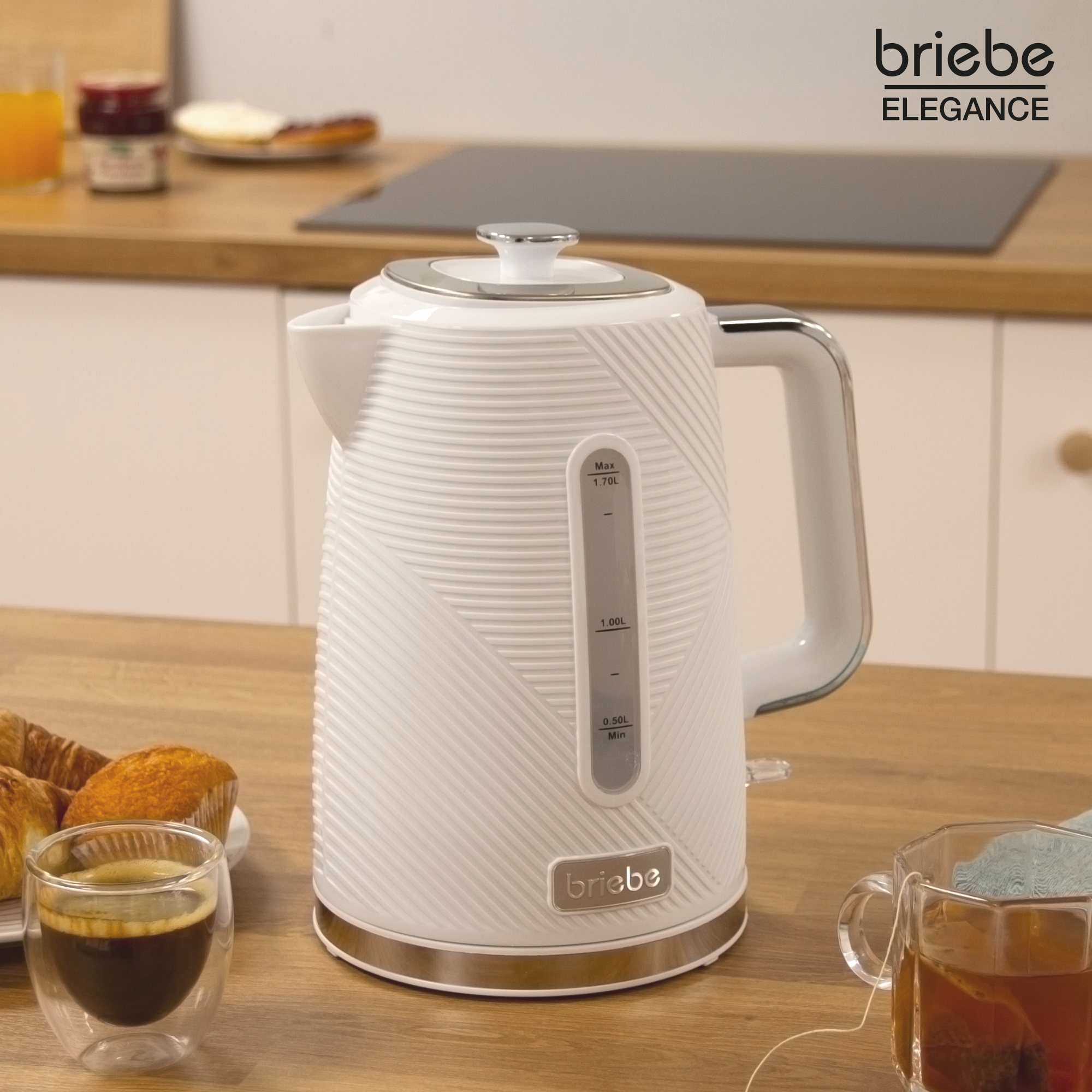 Hervidor blanco sobre mesa de madera, con café, pasteles y taza de té. Marca Briebe.