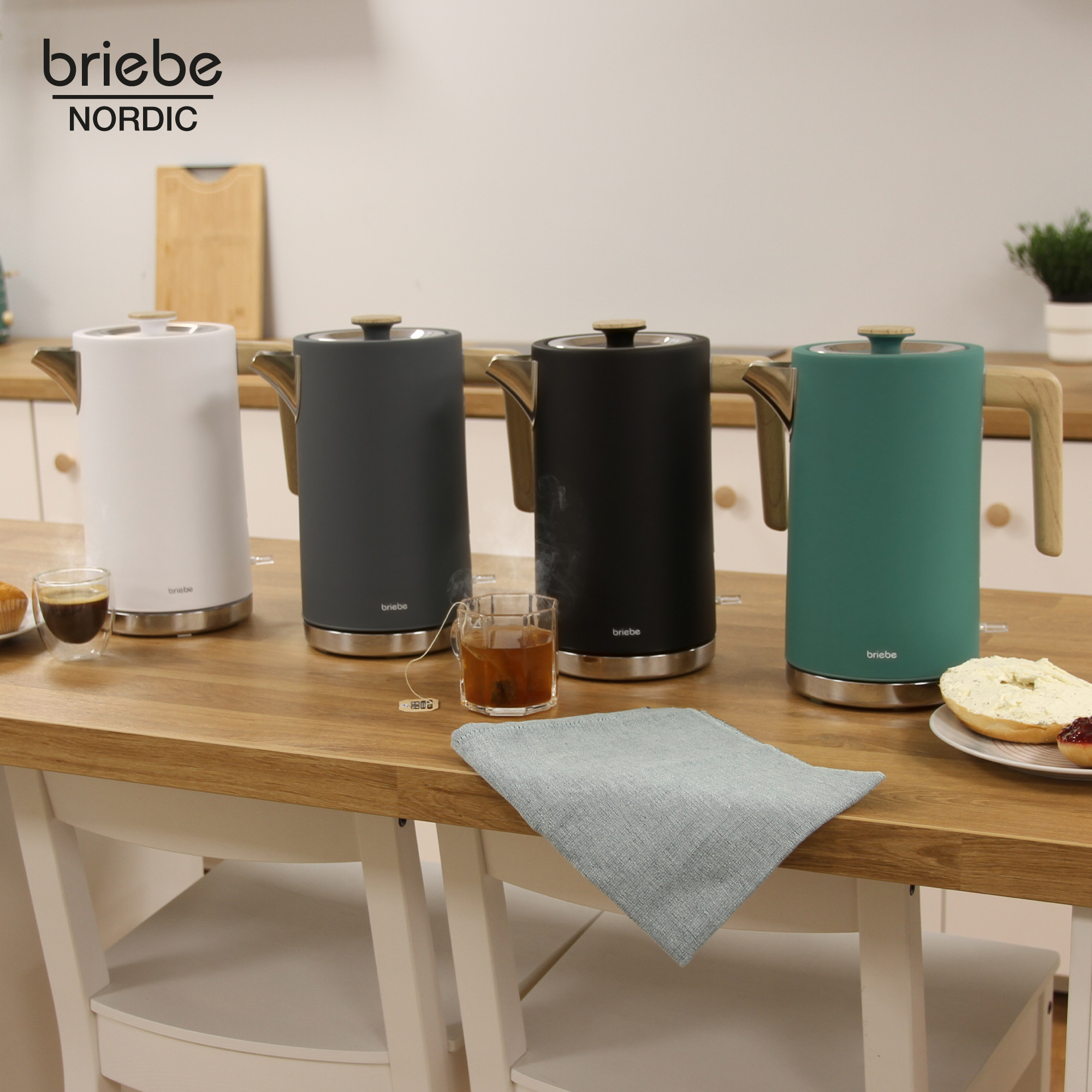 Cuatro hervidores eléctricos en diferentes colores sobre una mesa de madera con té y bollería.