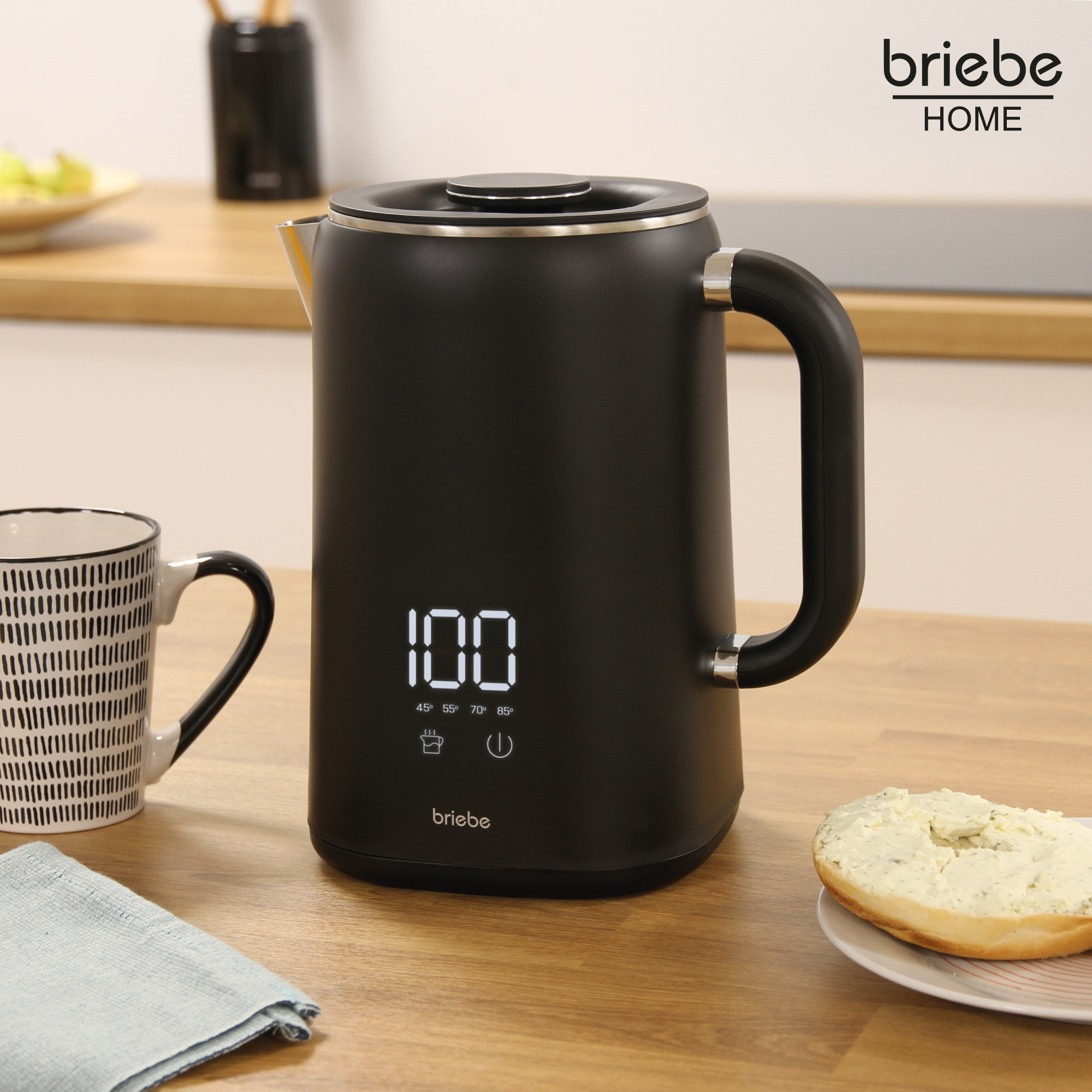 Schwarzer Wasserkocher auf Holztisch. Tasse und Bagel mit Frischkäse sind auch da.