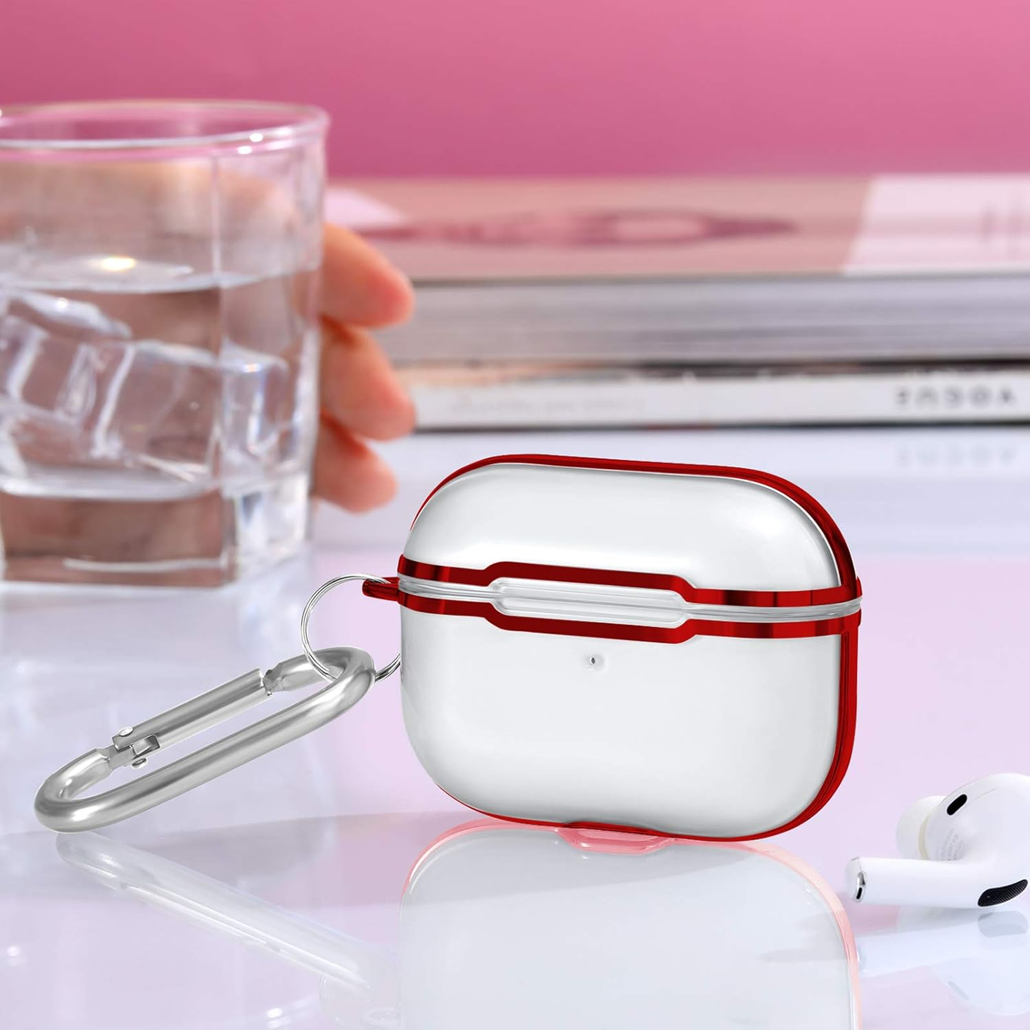 Weiß-rotes AirPod-Etui mit silbernem Karabiner. Ein Glas Wasser und ein Buch sind im Hintergrund.