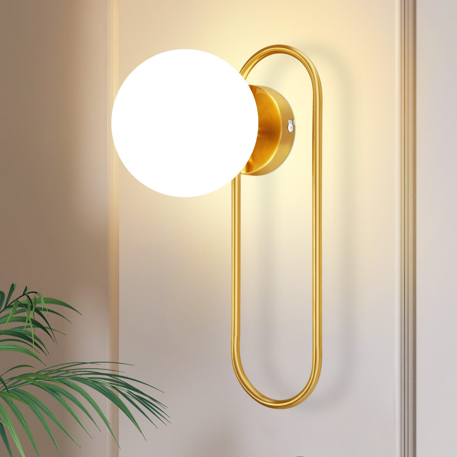 Lampada da parete dorata con luce bianca rotonda e cornice dorata curva, accanto a una pianta.