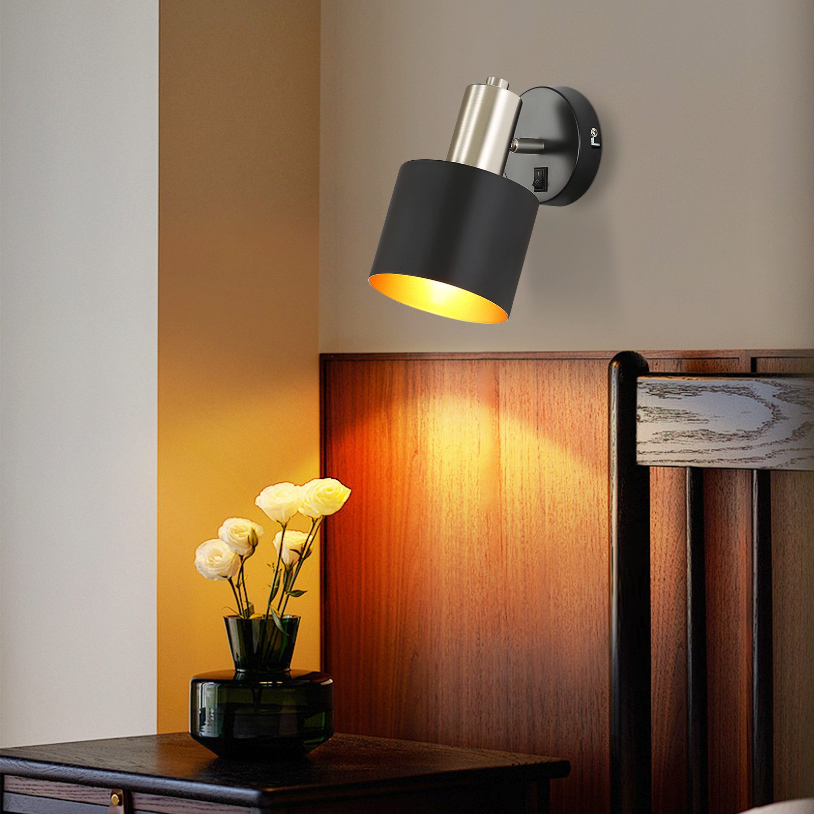 Lampada da parete nera e oro, che illumina un muro. Vaso con fiori bianchi sul tavolo.