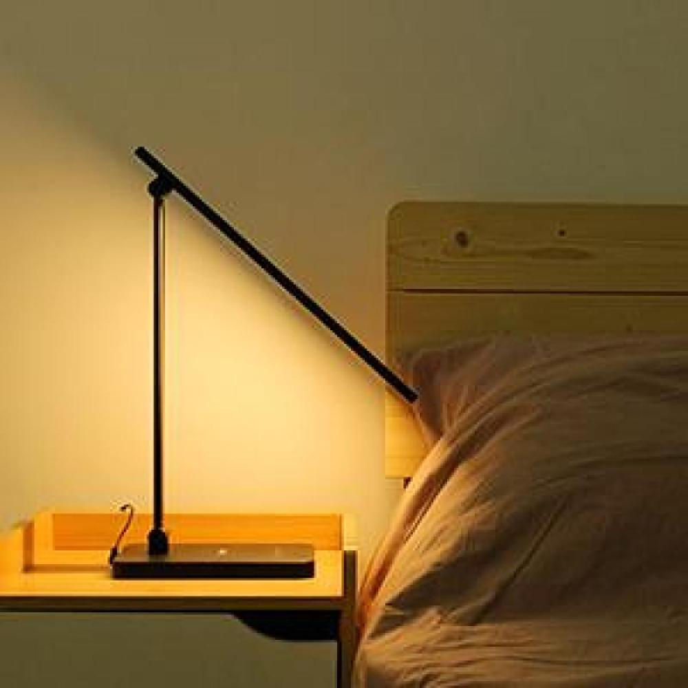 Eine schwarze Schreibtischlampe beleuchtet einen Nachttisch und ein Bett. Die Lampe steht auf einem Holztisch.